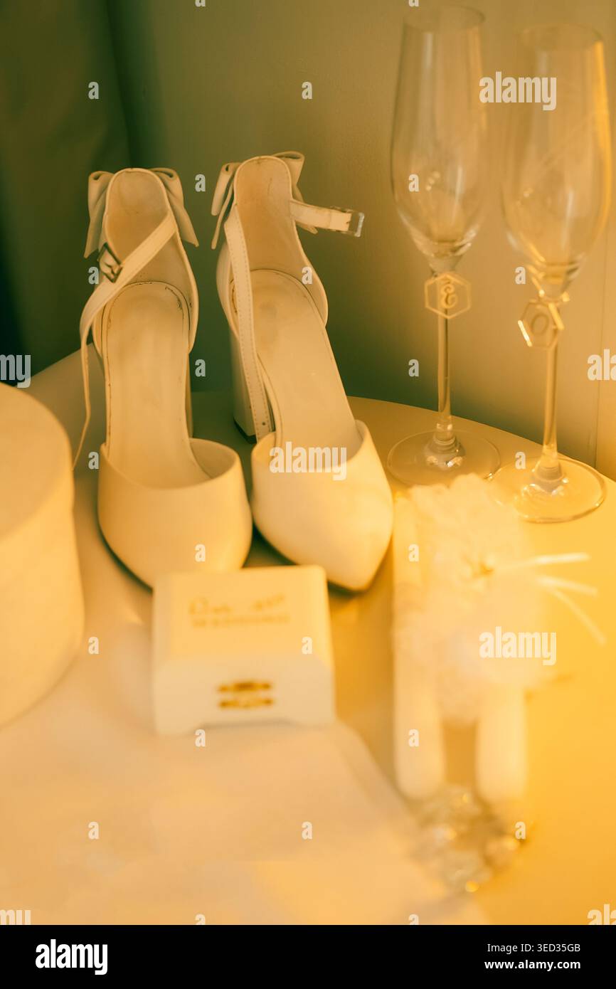 Elegant white heels prepared for wedding celebration. White high heels sit beside two champagne glasses, small gift box, and decorative item. setting Stock Photo