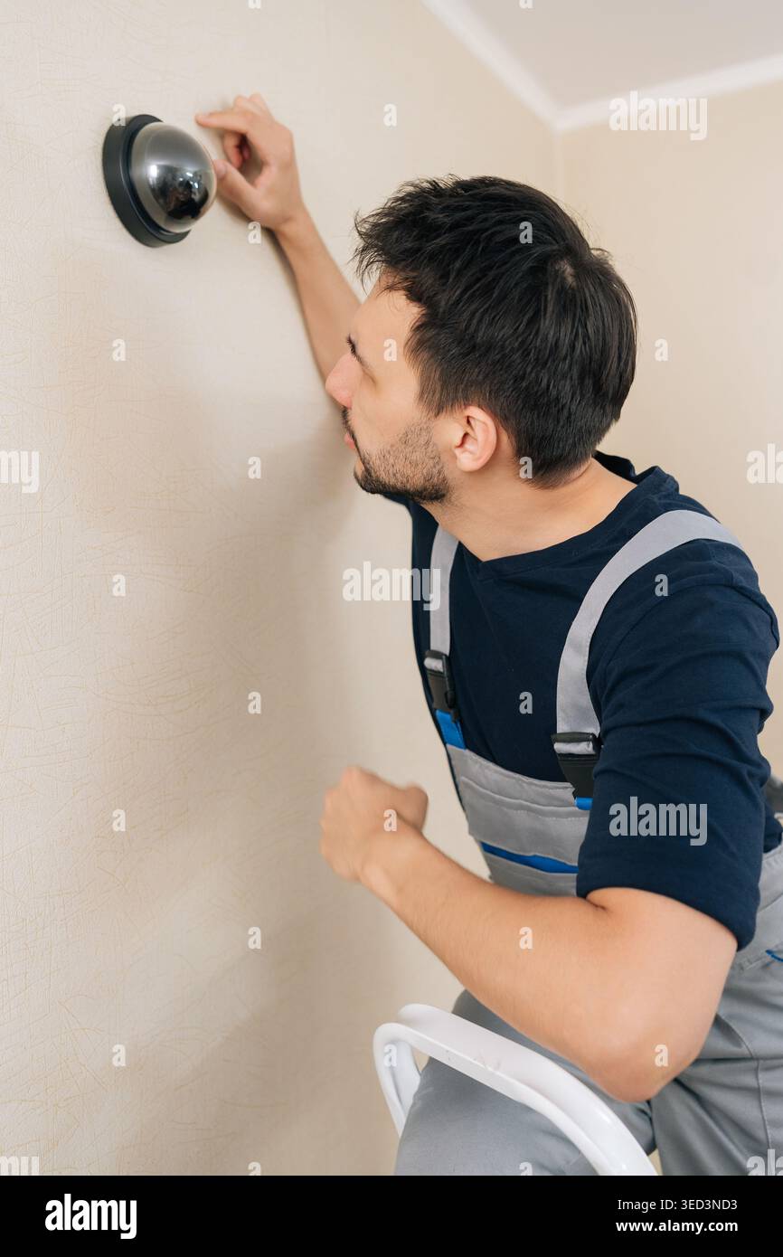 Professional handyman in uniform installs home security system. Male technician sets up black dome surveillance camera on wall for safety protection Stock Photo