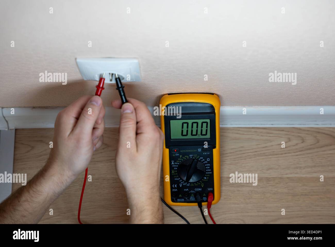 Hands Testing Wall Outlet With Multimeter, DIY Homeowner Performing Safety Check Near Baseboard, Probes Inserted Into Faceplate, Digital Meter Reading Stock Photo