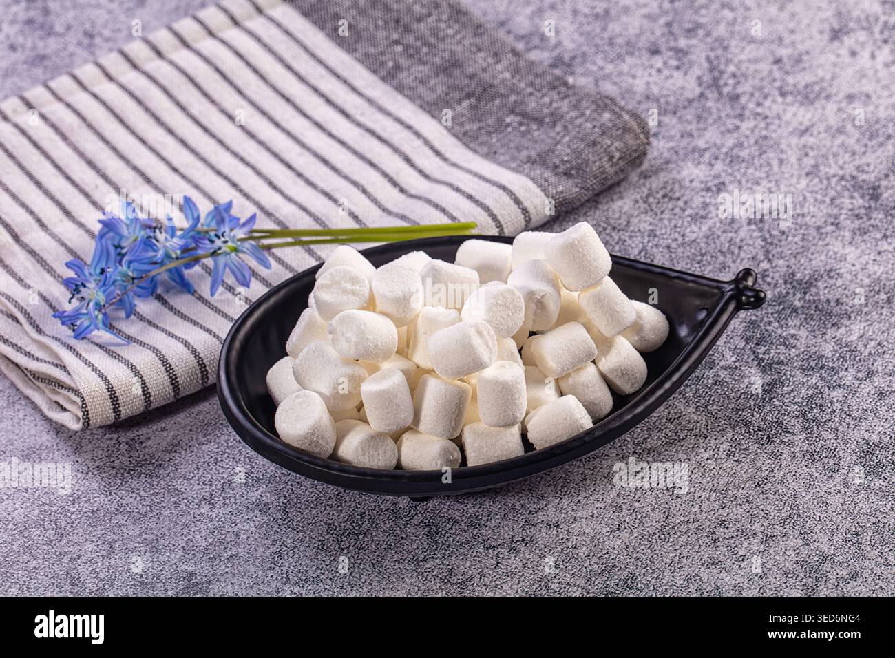 Fluffy white mini marshmallows fill black bowl. Striped grey, white napkin, delicate blue spring flowers rest beside Stock Photo