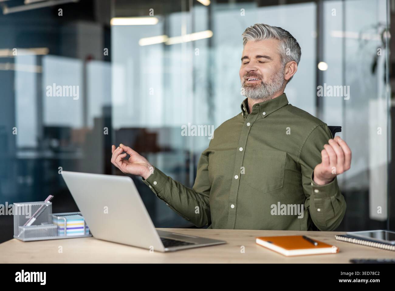Mature businessman closing eyes and meditating at his desk in a blur-background office, practicing mindfulness and stress relief during a work break by finding zen moment Stock Photo