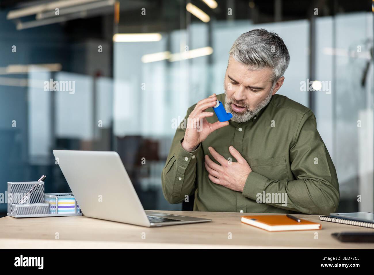 Mature man suffering from an asthma attack and breathing difficulties while sitting at his office desk, using an inhaler for relief and urgently needing treatment Stock Photo