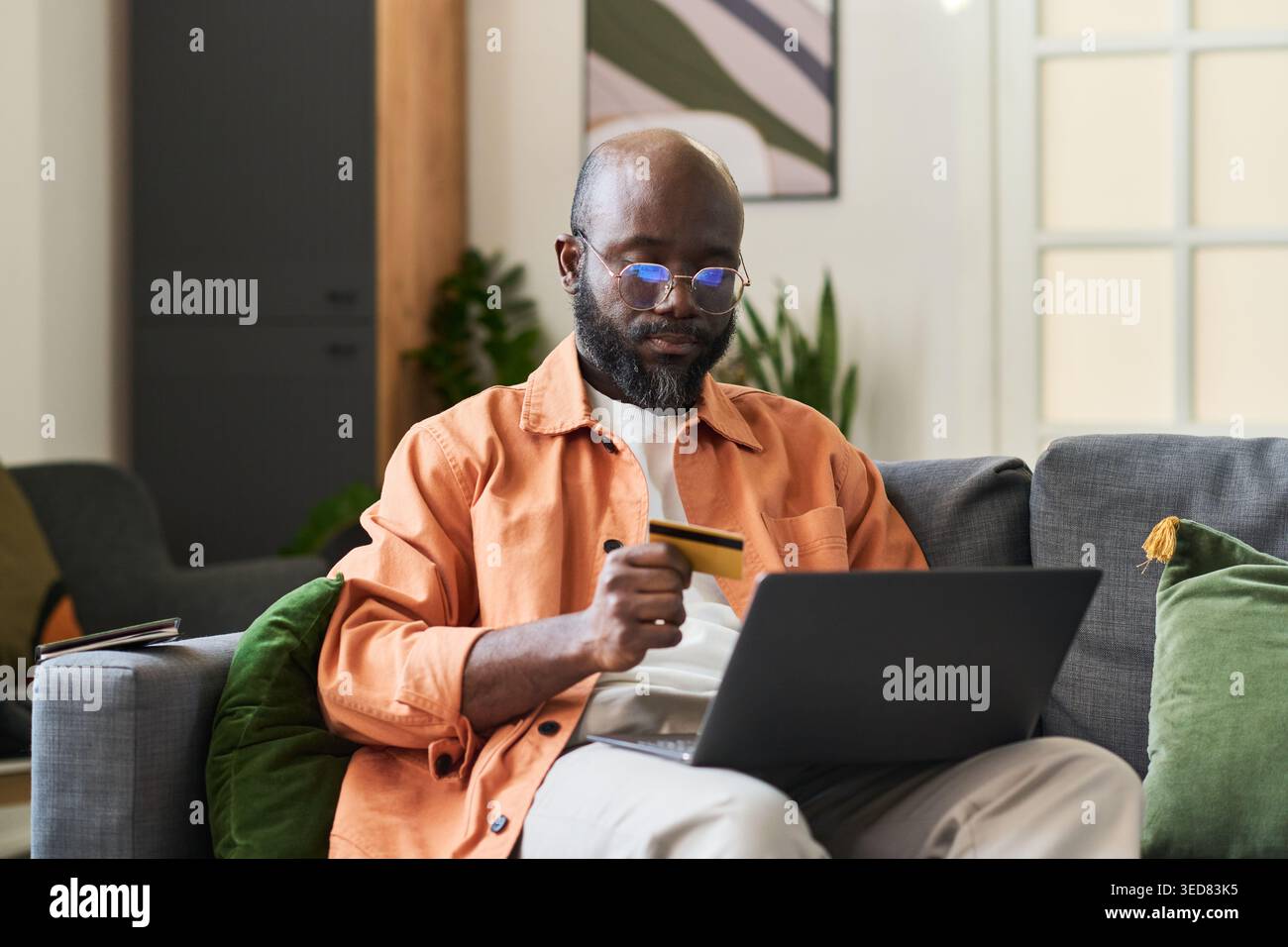 Medium shot of Black middle aged man holding credit card and using laptop while booking trip from home, planning travel purchase. Useful for tourism marketing, online payment Stock Photo