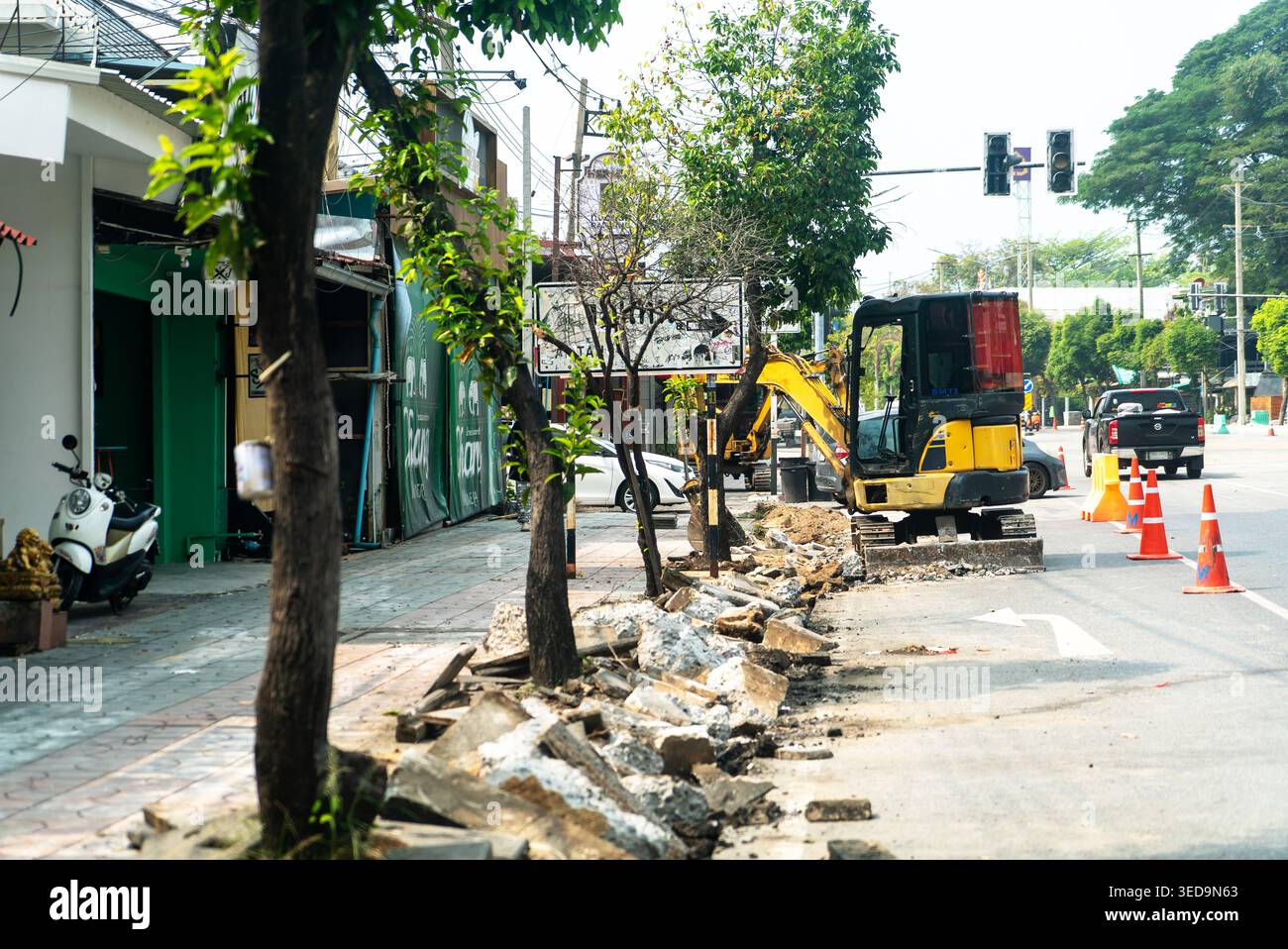 Construction work is underway on an urban roadside area, featuring machinery, traffic cones, and trees, showcasing the bustling environment of city ma Stock Photo