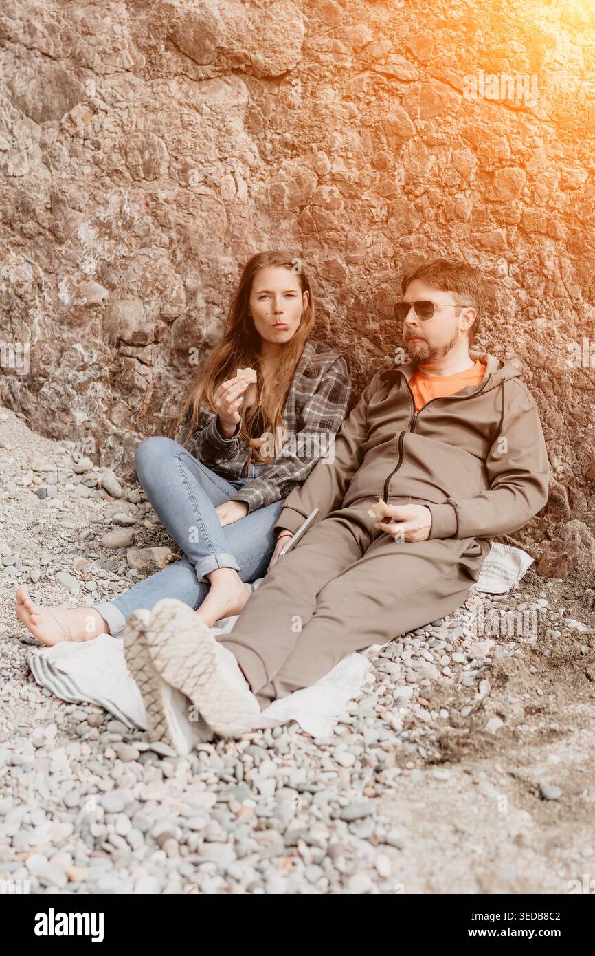 Couple beach picnic enjoying snacks while sitting on rocky coastline near a rock wall under sunlight Stock Photo