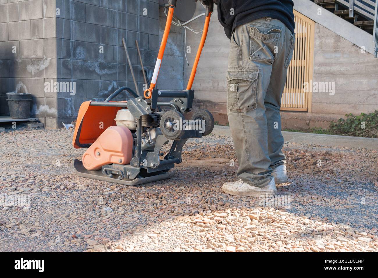 Worker operates a vibratory plate compactor on a gravel surface at a construction site. The machine compacts the ground to create a stable base for Stock Photo