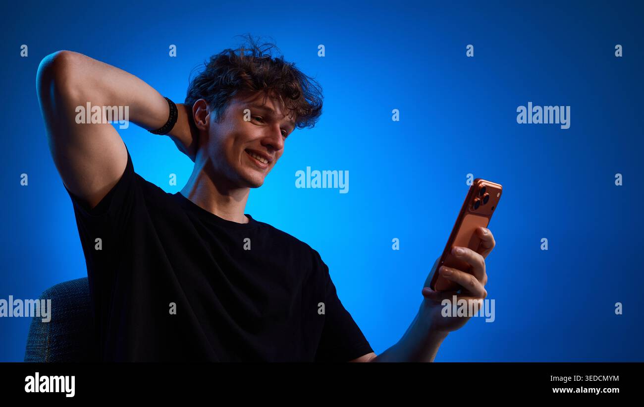 Smiling young man reading smartphone message in blue studio light Stock Photo
