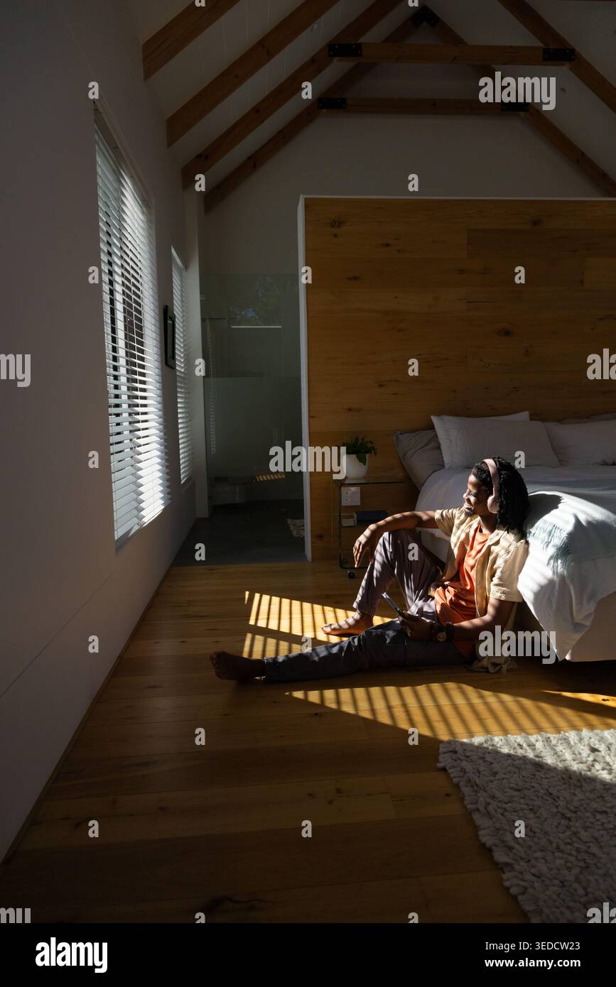 Adult African man sitting on wood floor next to bed holding smartphone, headphones on, copy space Stock Photo
