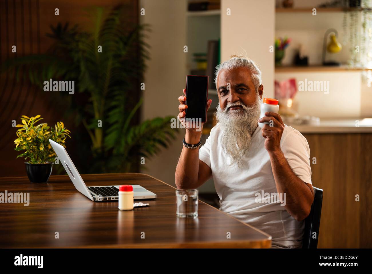Indian mid age man checking medicine ingredients online holding pills bottle while speaking on phone and using laptop at table in modern kitchen inter Stock Photo