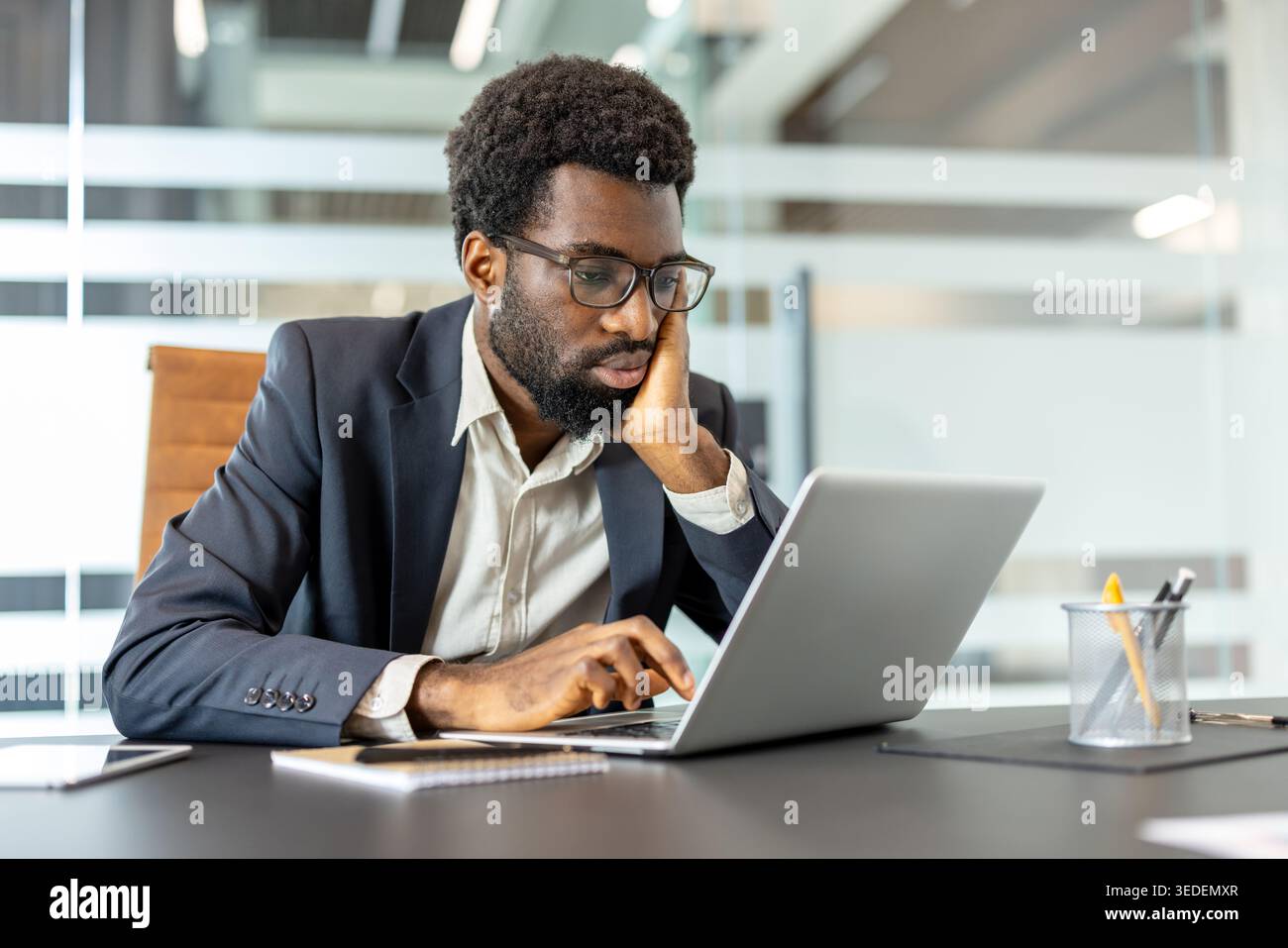Young businessman wearing glasses and a suit, sitting at a desk with his hand supporting his chin, deeply concentrating on his laptop screen in a modern corporate office setting Stock Photo