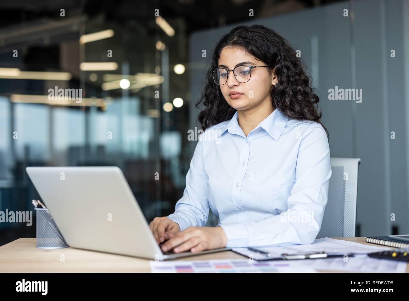 Young indian businesswoman wearing glasses and a blue shirt, concentrating while typing on a laptop computer at a desk in a modern corporate office environment Stock Photo