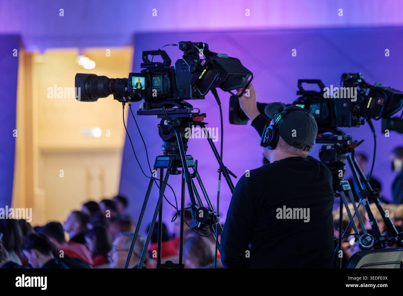 Cameraman operates professional video camera on tripod during live conference, colorful stage lighting and audience. Stock Photo