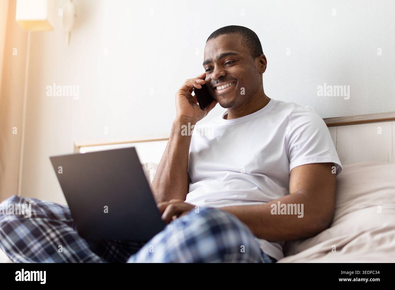 African american man talking on smartphone while using laptop on bed Stock Photo