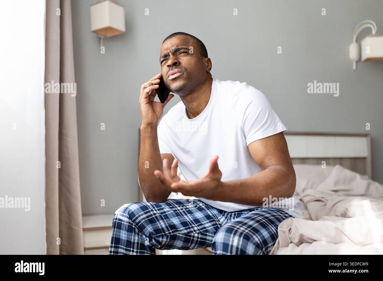 Black man talking on smartphone while sitting on bed, making expressive gesture Stock Photo