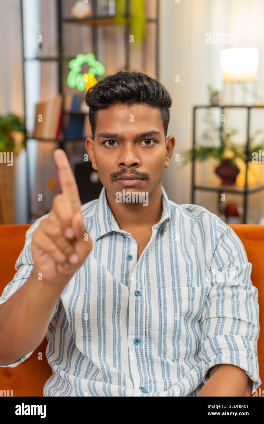 Indian man at home waves finger no no to camera and shows refusal and disagreement with stern mood. Arabian guy on sofa shakes head denies request points finger and shows unhappy sad reject reaction Stock Photo