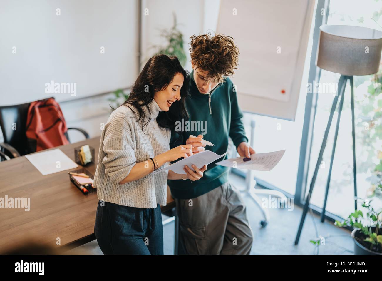 Two students collaborating over documents in a bright modern classroom while smiling and discussing Stock Photo