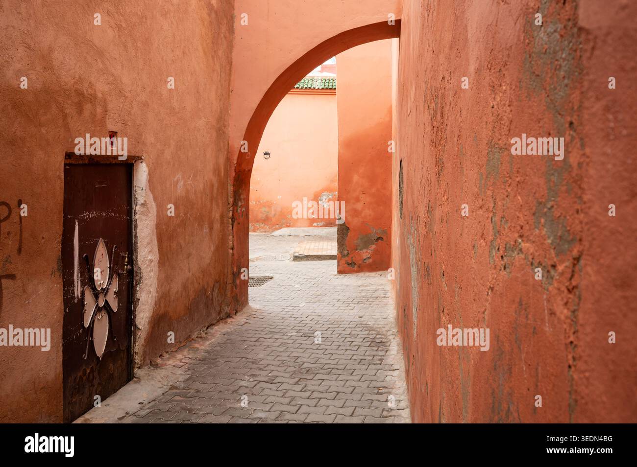 Winding back alley in the Marrakech medina framed by an arched passage, orange textured walls and a weathered wooden door. Atmosphere feels historic a Stock Photo