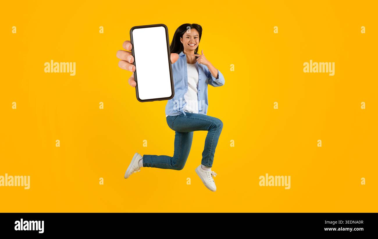 Woman Jumps and Shows Smartphone in Bright Studio With Yellow Background Stock Photo