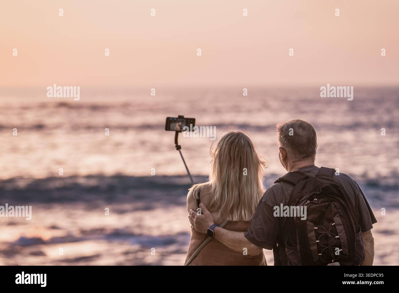 Mature couple taking selfie on the beach at sunset with smartphone and selfie stick, standing together near the sea while looking at the ocean. Warm e Stock Photo