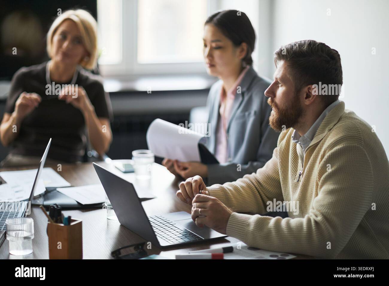 Businessman discussing strategy with multiethnic colleagues during office meeting, using laptops and documents. Ideal for teamwork, leadership, planning, corporate communication Stock Photo