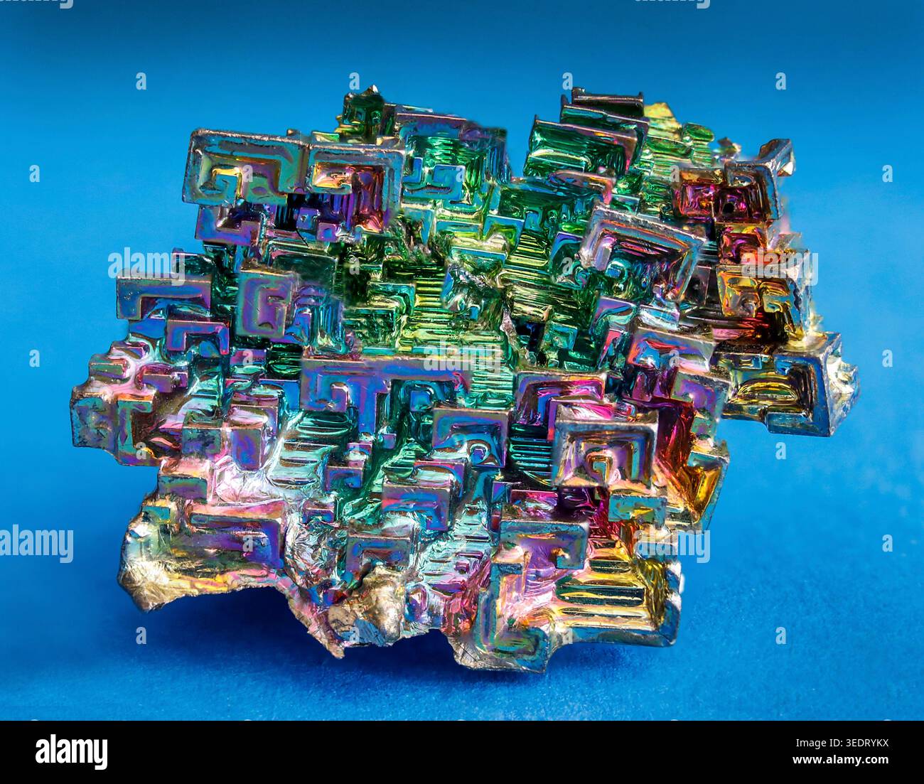 Iridescent bismuth crystal with mesmerizing geometric hopper formations, shimmering in vivid rainbow metallic colors against blue. Stock Photo