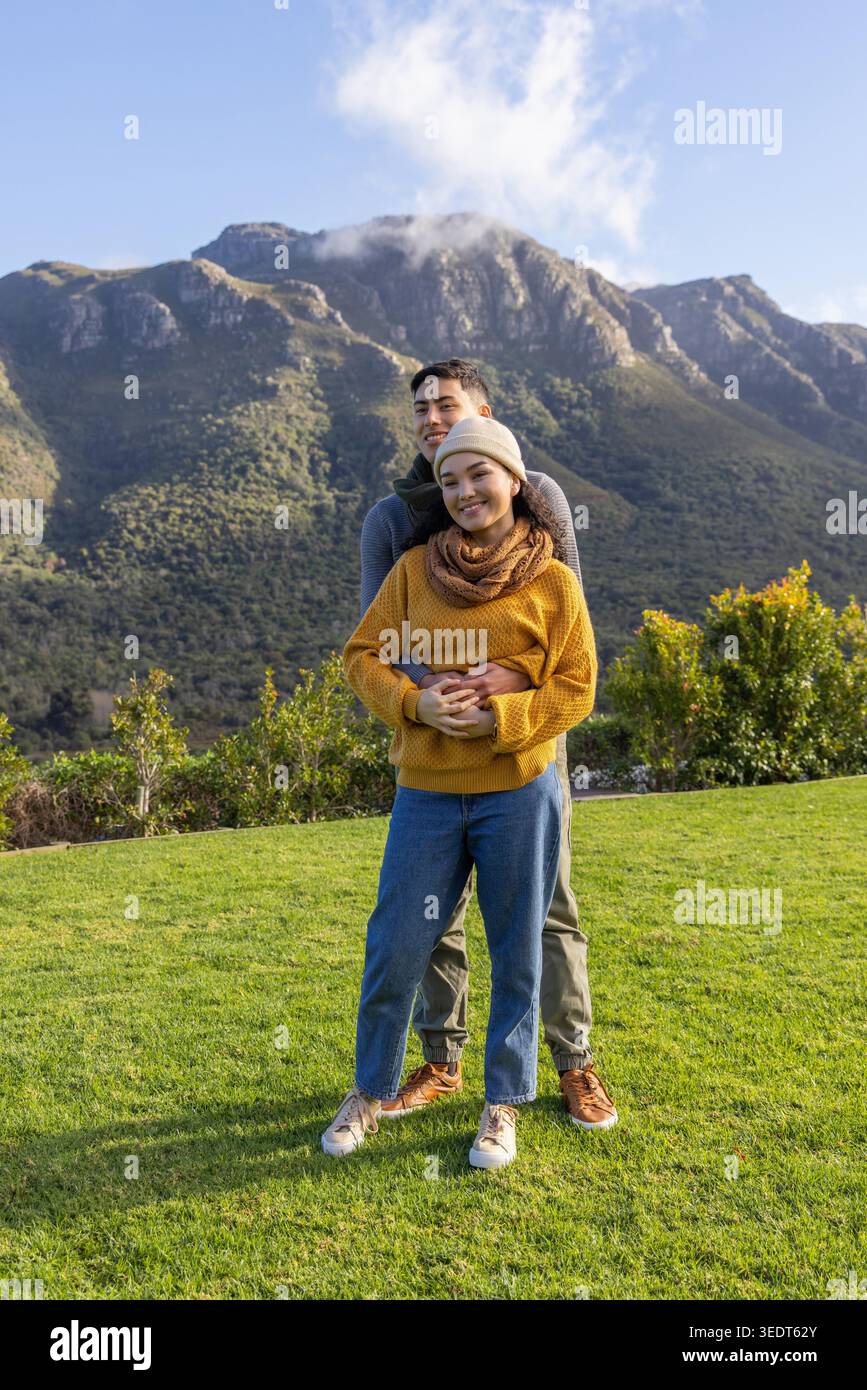Asian couple standing embracing on lawn at mountain foothills, wearing knit sweater beanie scarf. Romance, outdoors, pair, alpine, ridge, scenery, greenery Stock Photo