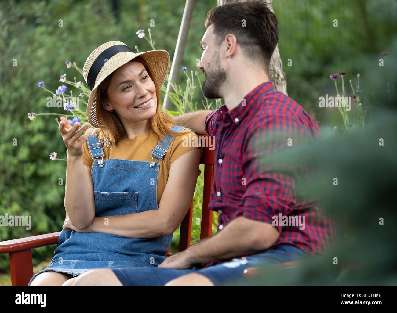Young man and woman sit on a bench in a garden. They are smiling and enjoying a conversation surrounded by plants and flowers. Stock Photo