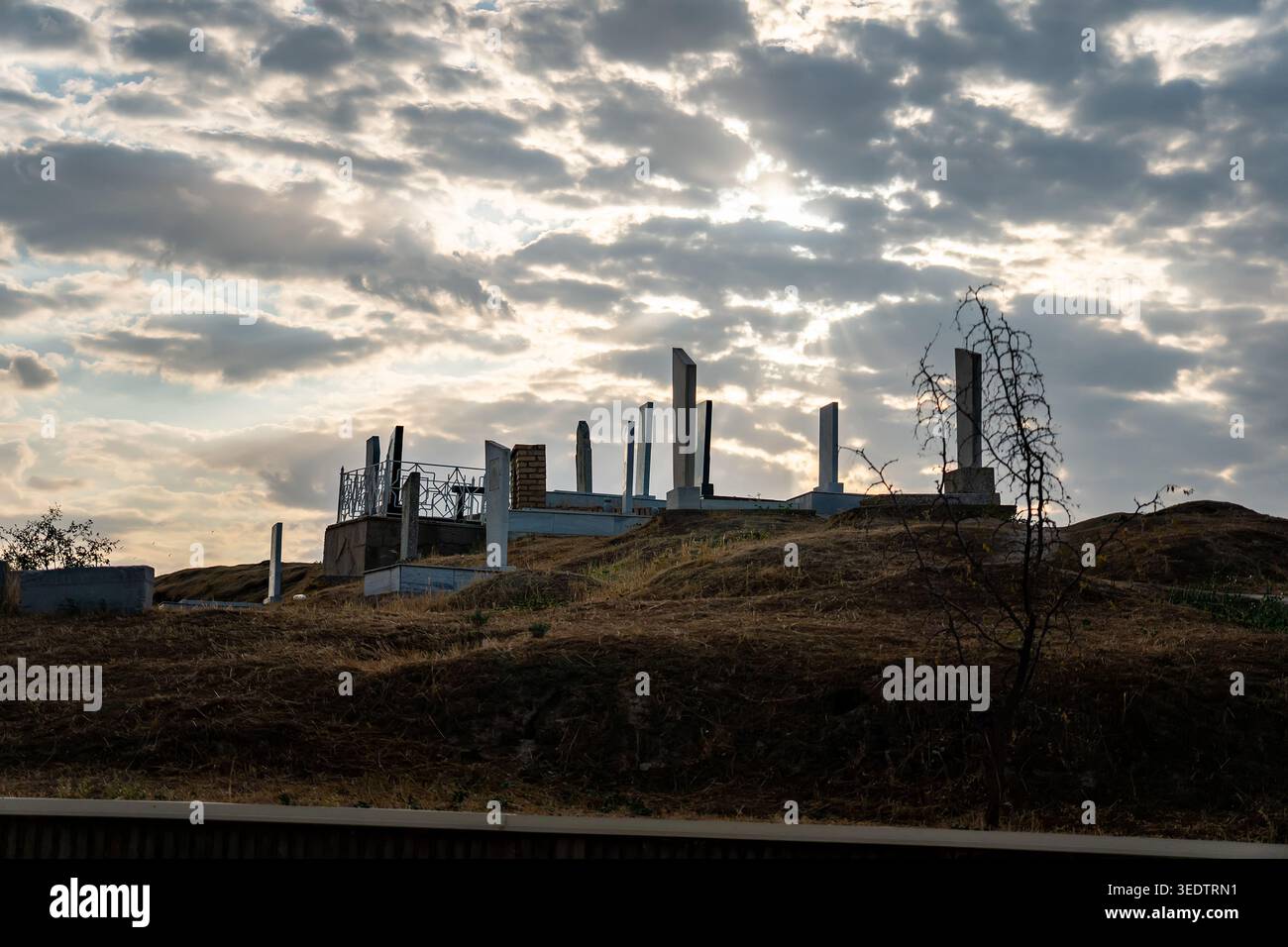Dramatic sunset over old cemetery with ancient stone stelae and burial mounds on a hill. Bright sun rays breaking through dramatic clouds create power Stock Photo