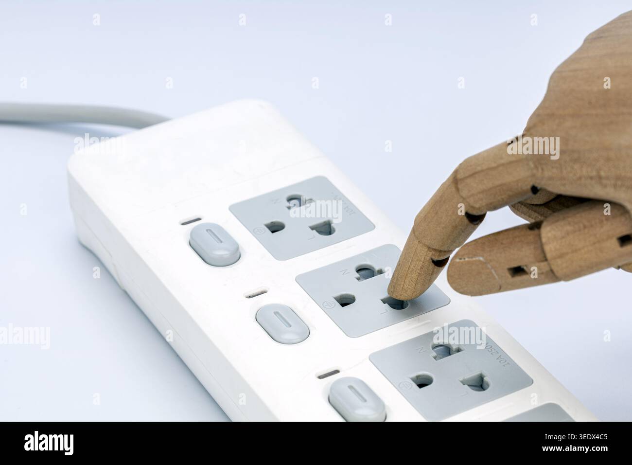 A wooden jointed mannequin hand pokes its finger directly into a live electrical outlet on a white multi-socket power strip. This striking image power Stock Photo