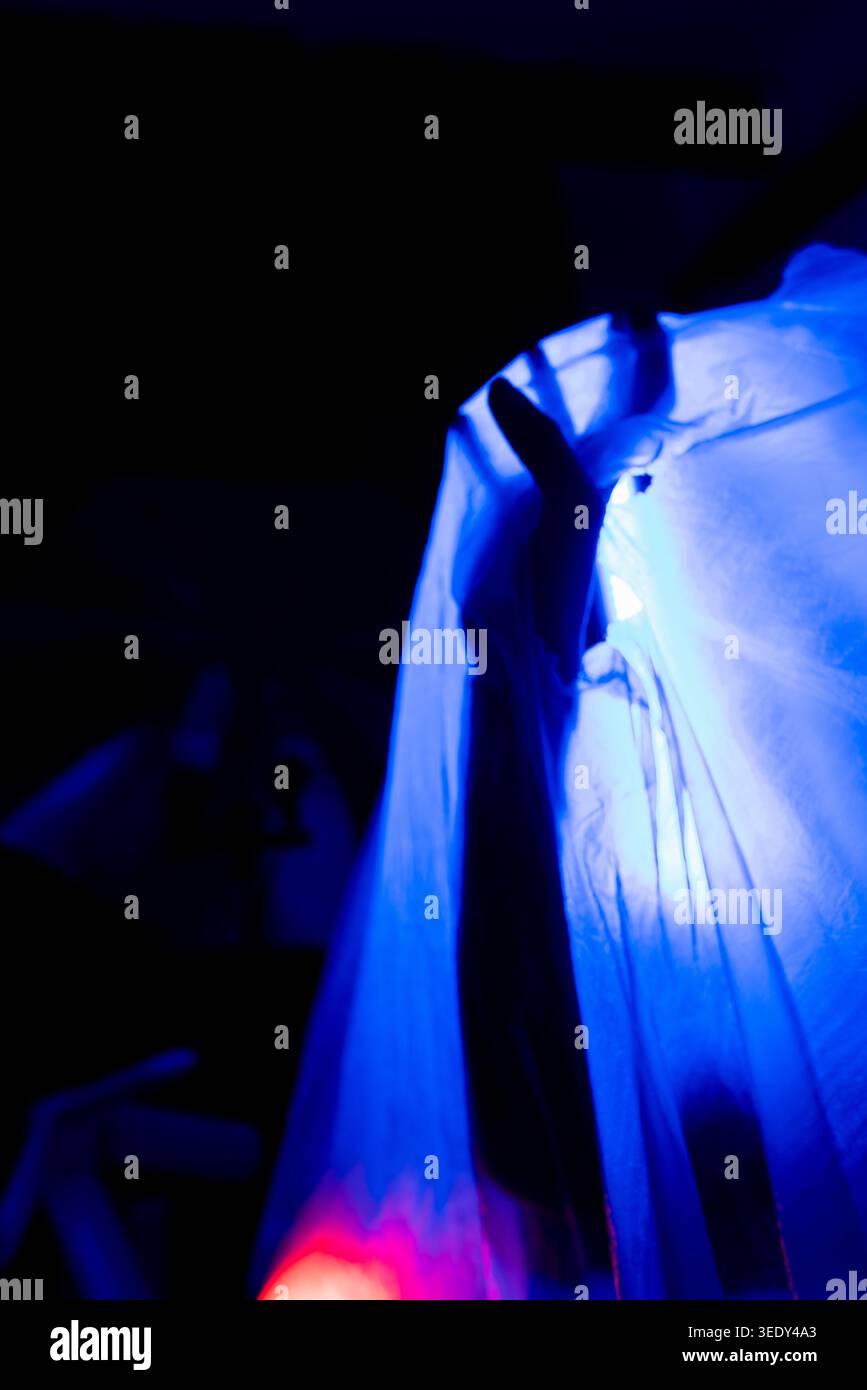 Minimal dark silhouette of a hand pressing through white translucent fabric under blue neon lighting. Conceptual horror and emotional isolation scene Stock Photo