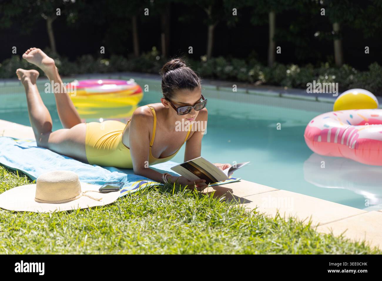 Woman lying on blue towel at backyard pool edge, wearing yellow swimsuit, reading paperback Stock Photo