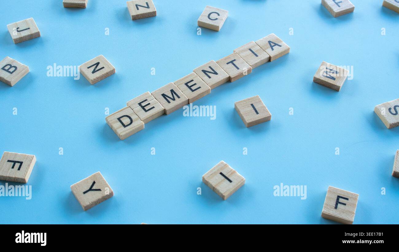 High angle view of wooden tiles spelling the word dementia at a diagonal angle over a blue backdrop with mixed alphabet letters Stock Photo