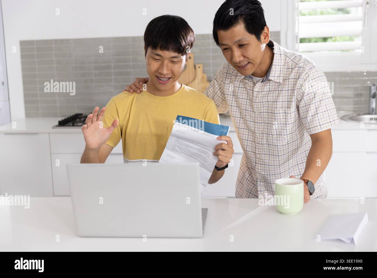 Asian family members reviewing documents on laptop at home kitchen with green mug and envelope. Duo, computer, papers, letter, cup, stock, interiors Stock Photo