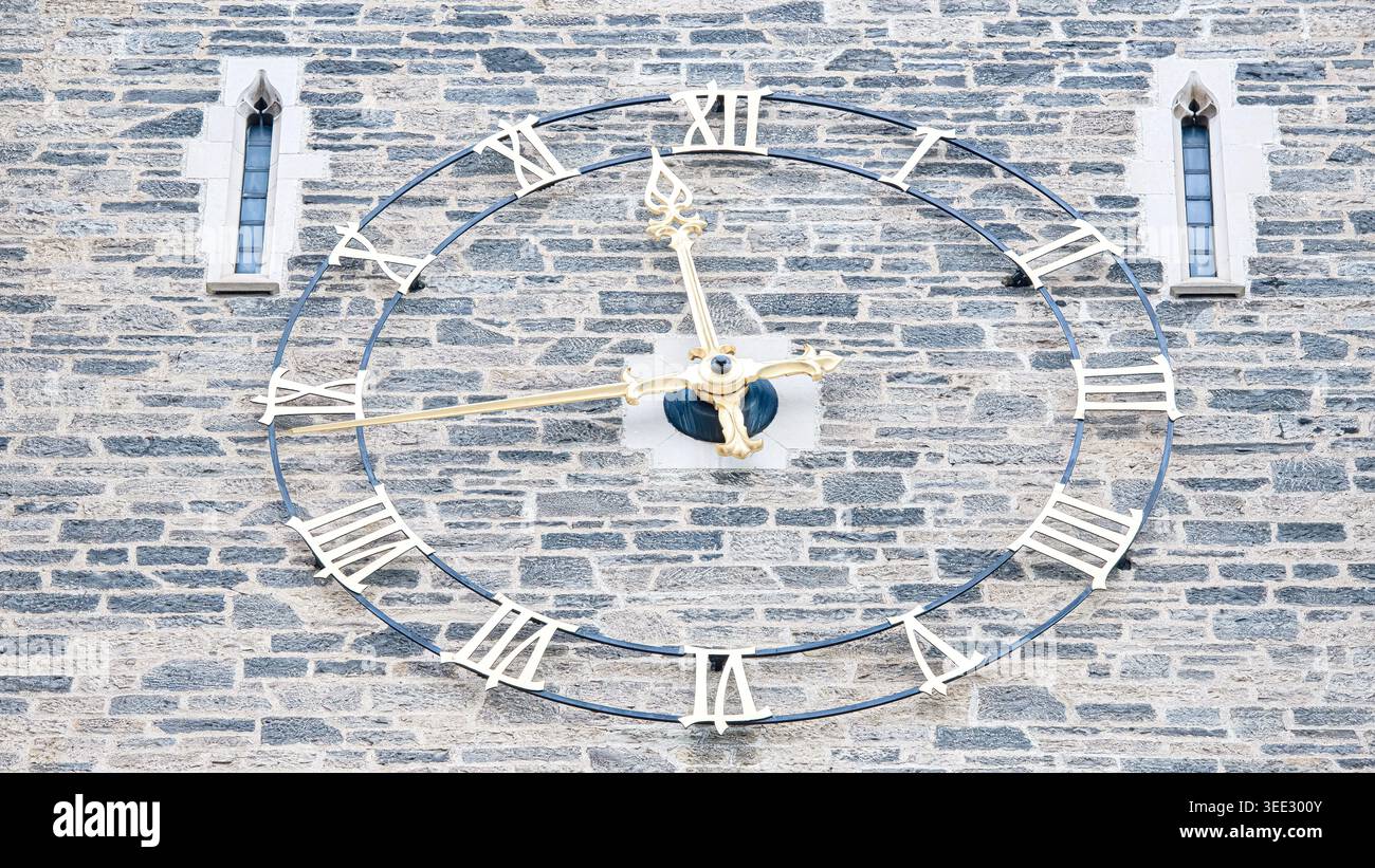 Close view of Soldiers Tower clock with golden hands and Roman numerals set against textured stone wall with narrow vertical windows. Stock Photo