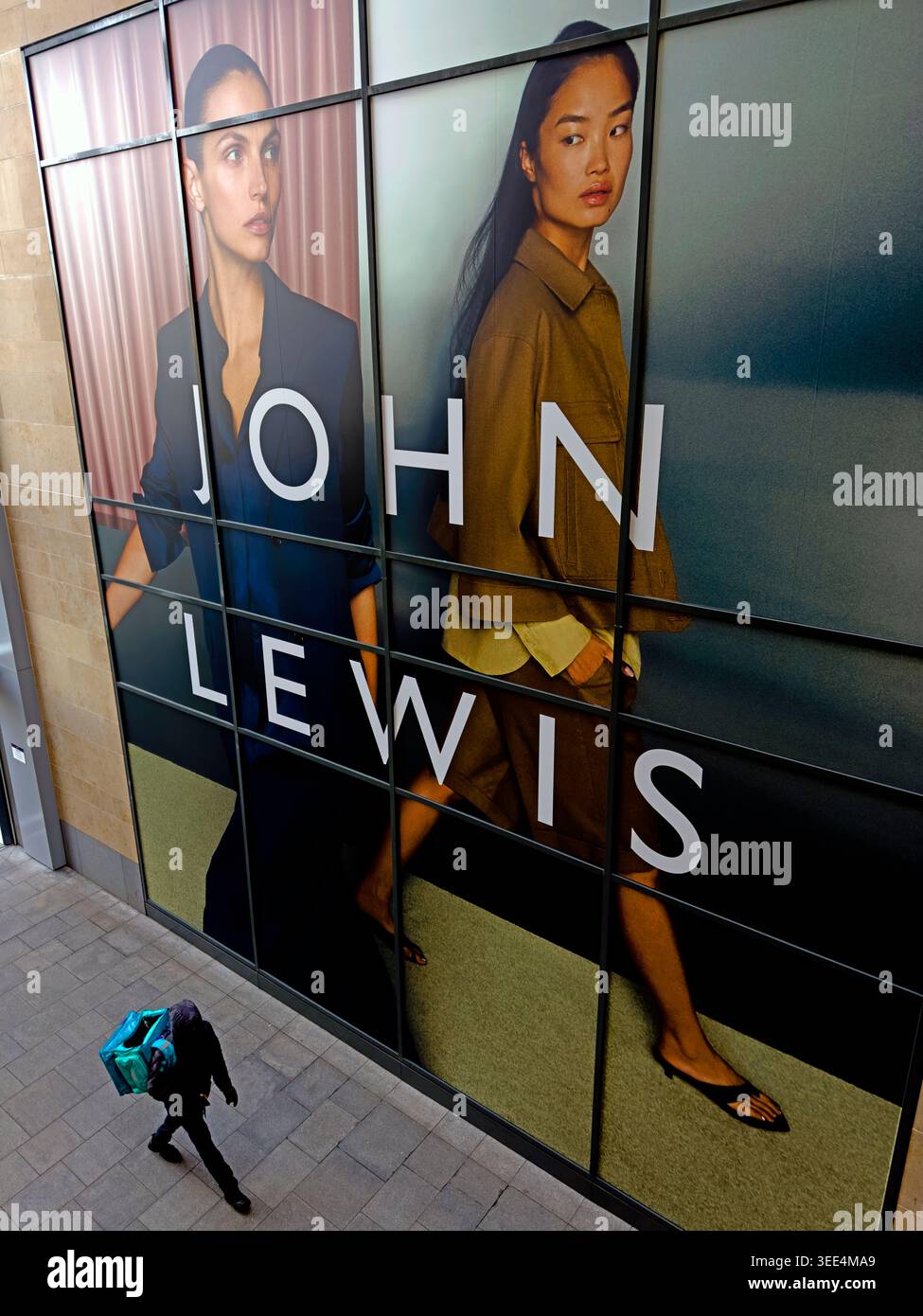 Food delivery rider passes below a large John Lewis fashion hoarding in Edinburgh city centre, highlighting gig work contrasts. Stock Photo