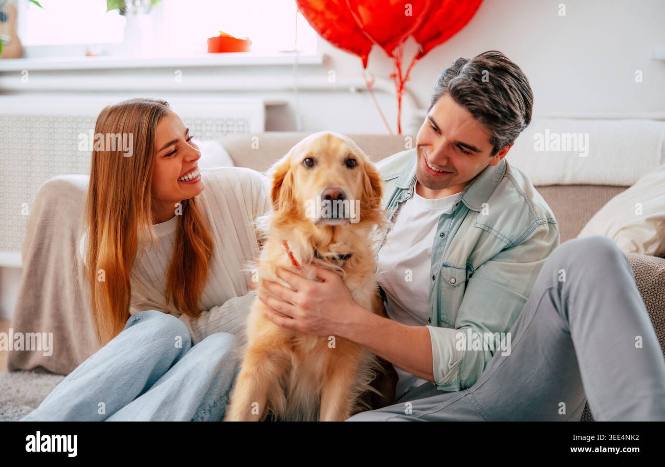 Happy young couple relaxing with their dog at home, smiling pet owners bonding with golden dog in cozy modern living room Stock Photo