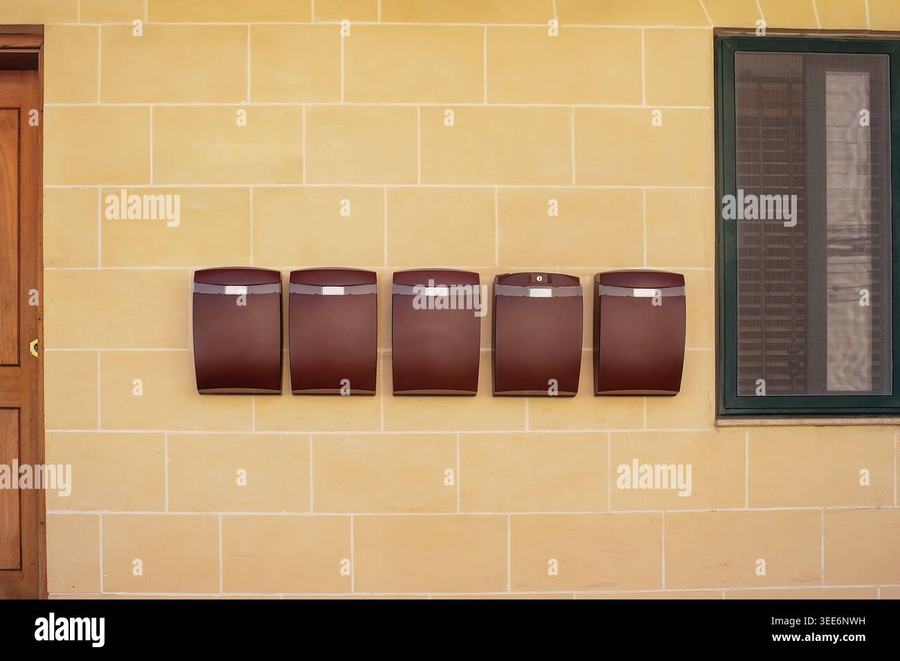 Several identical brown mailboxes on a yellow limestone wall between a door and a window. Concept of communication, postal service and house exterior. Stock Photo