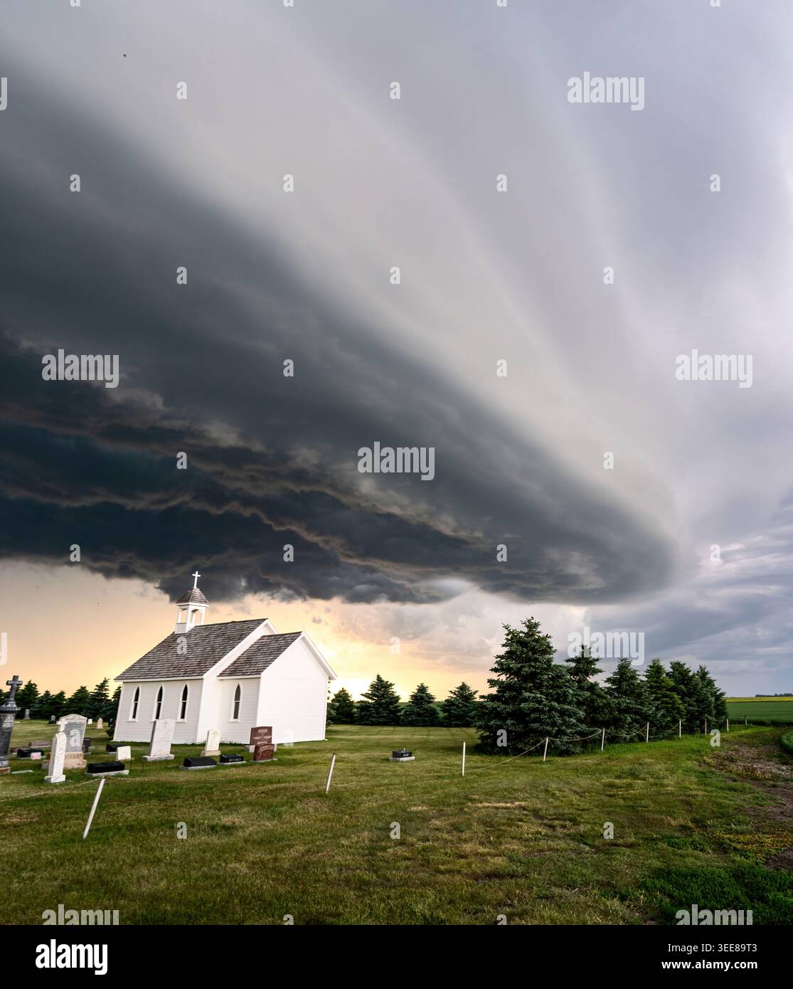 A serene white chapel stands in a graveyard beneath a vast, ominous shelf cloud formation during twilight Stock Photo
