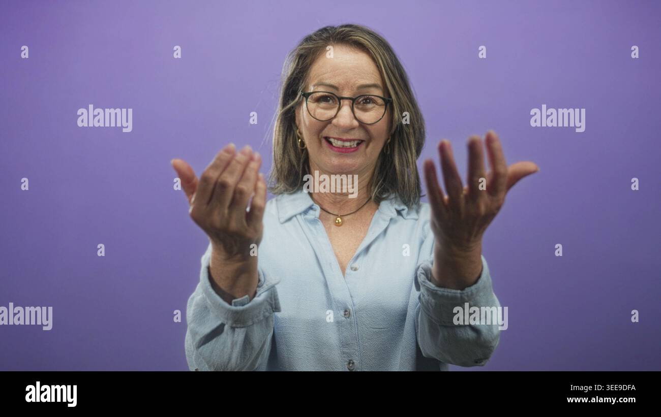 Hispanic woman with grey hair beckons with both hands toward camera in a studio wearing glasses and a light blue shirt; friendly invitation. Stock Photo