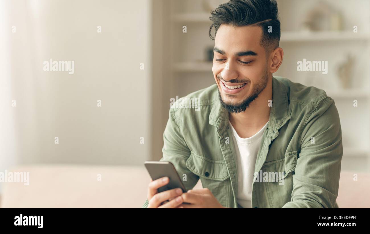Young Man Smiles While Using Smartphone at Home in Casual Setting Stock Photo
