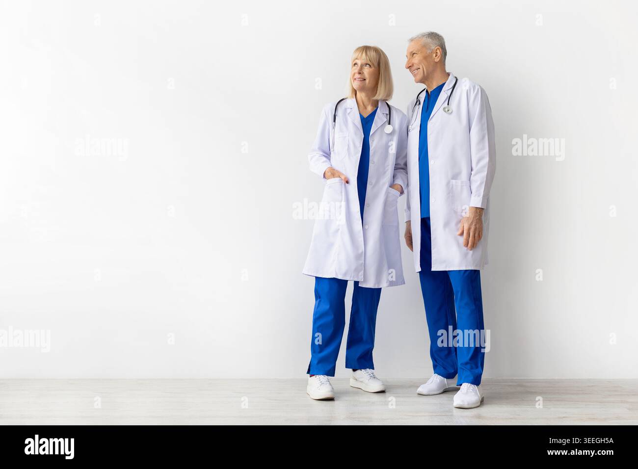 Two mature doctors in white coats and blue scrubs on white background Stock Photo