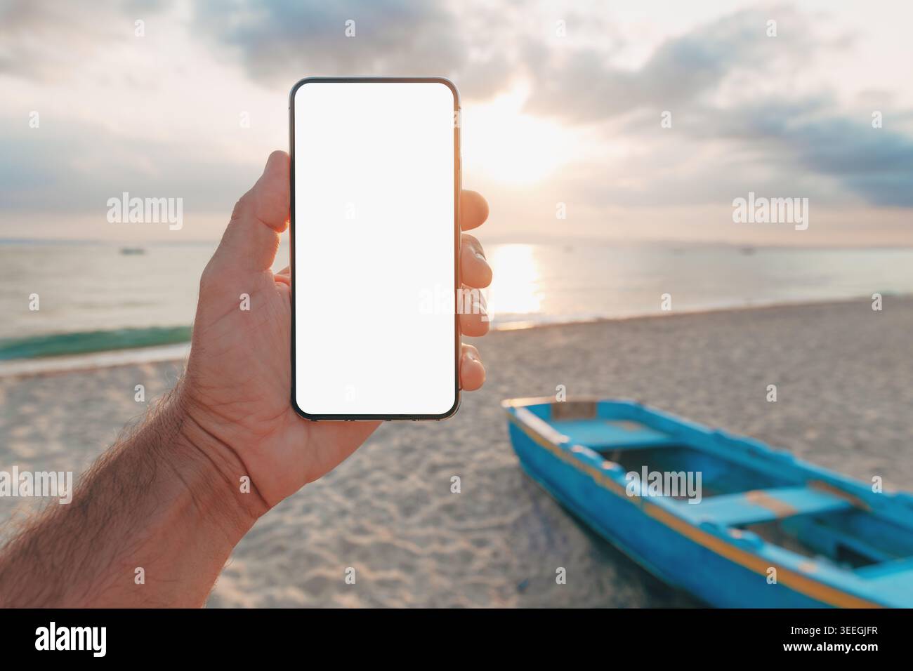 Hand holding smartphone with blank screen at beach with small blue boat and sunset in background, outdoor app mockup or web design. Selective focus. Stock Photo