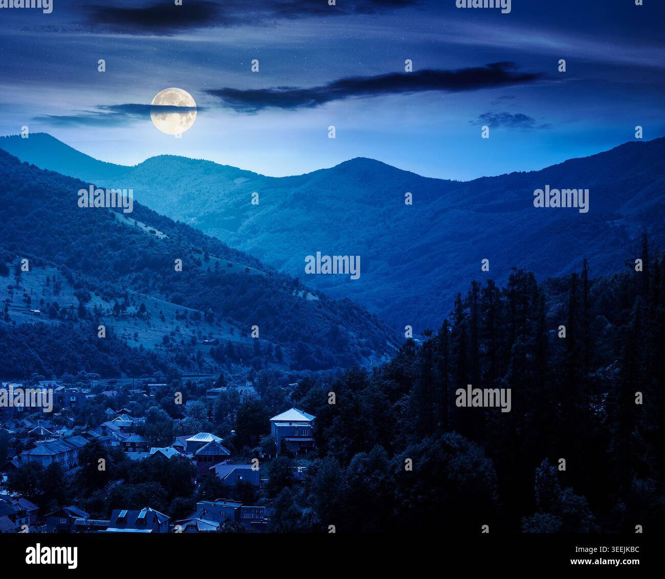 carpathian mountain landscape on summer night. full moon over view in to the valley of town near forested hill of mountain range. mysterious travel ba Stock Photo