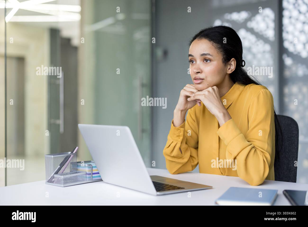 Professional woman in a yellow blouse seated at a modern office desk with laptop, deep in thought as she plans strategy, analyzes ideas and contemplates business solutions Stock Photo