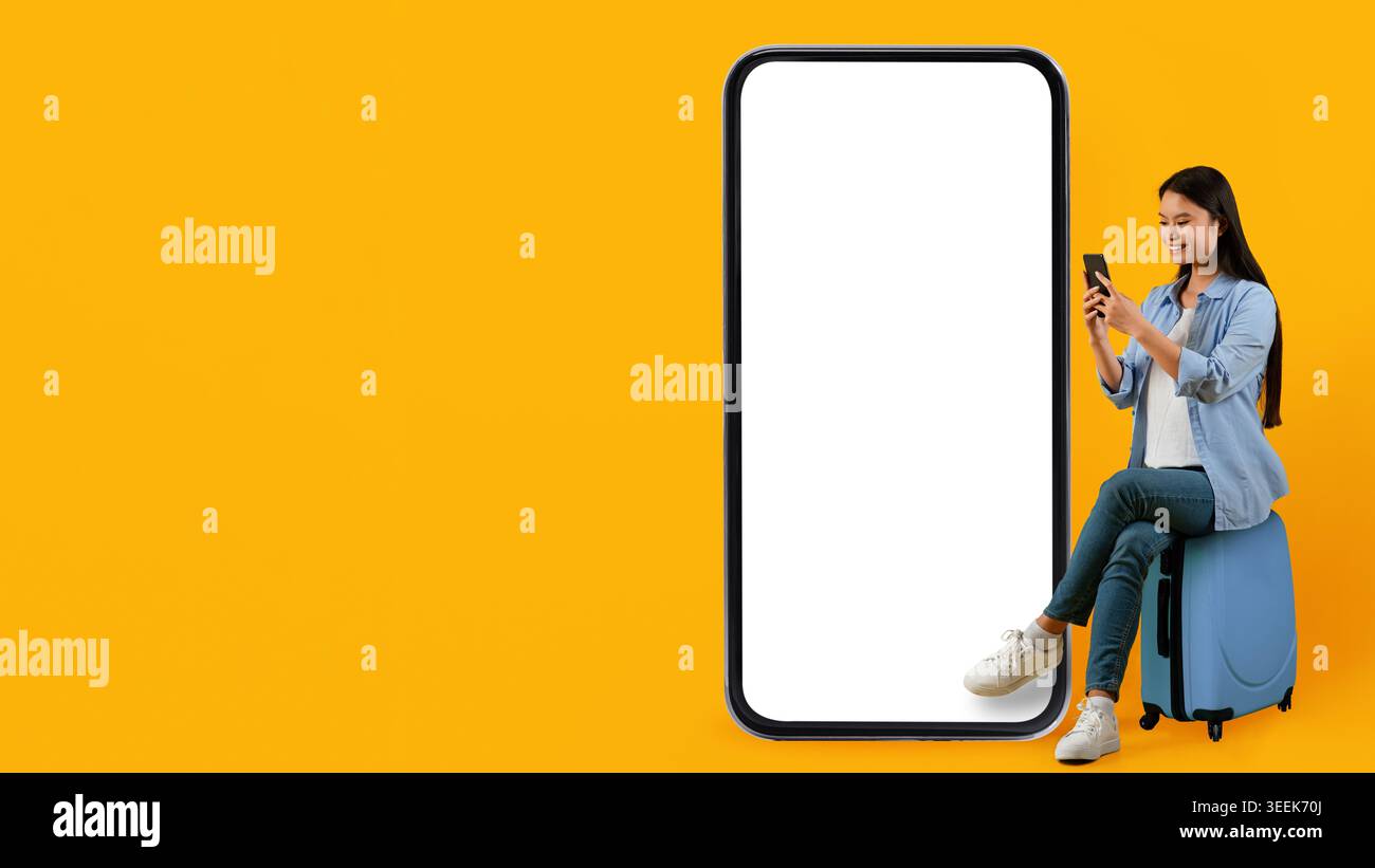 Woman Using Smartphone While Sitting on Suitcase Next to Large Blank Screen Stock Photo