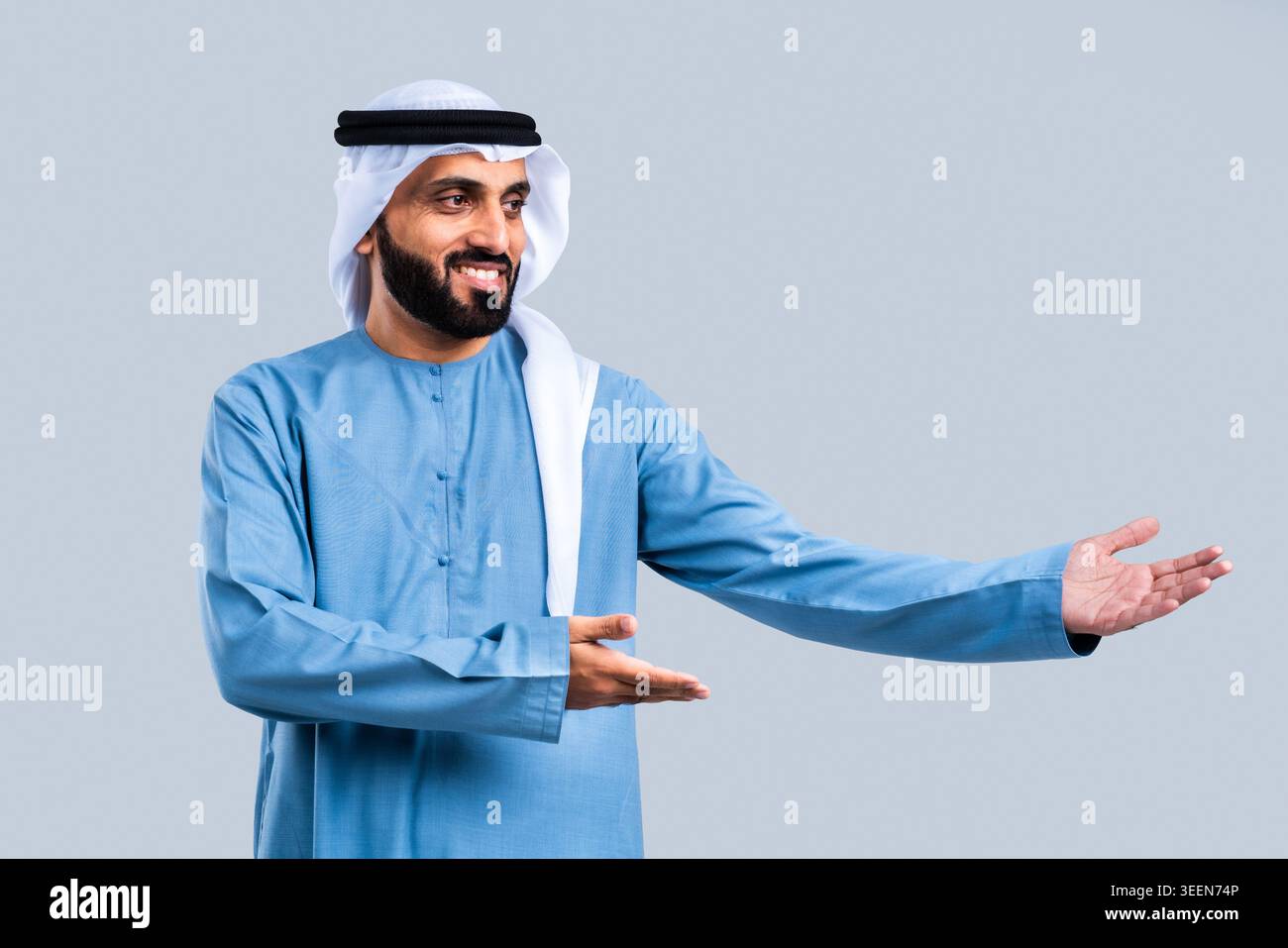 Emirati man wearing traditional kandura, ghutra, and agal, presenting with both hands while smiling against a plain background, conveying concepts of Stock Photo
