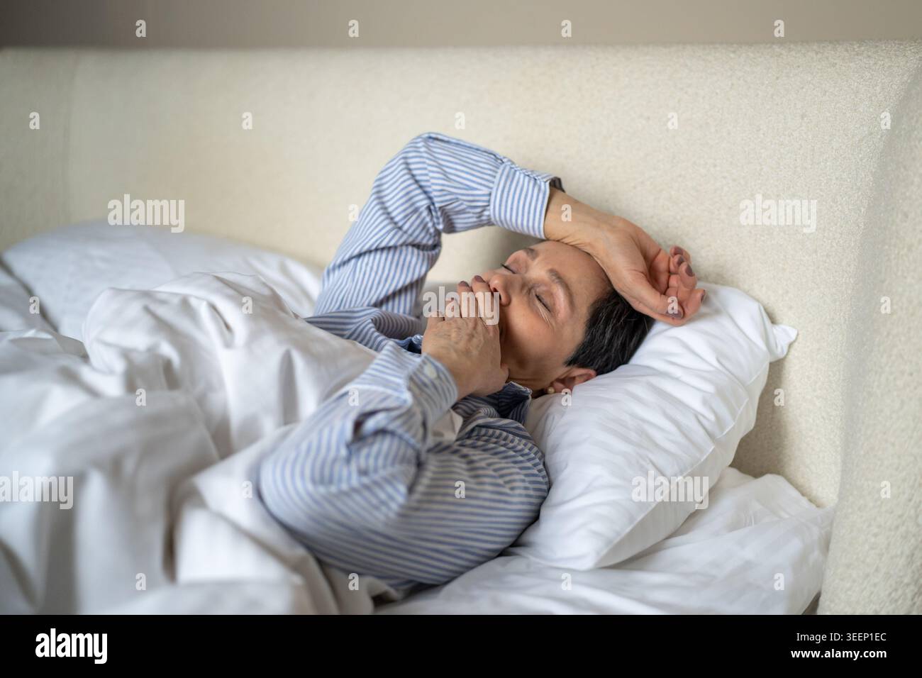 Dreamy senior woman comfortably lying in bed covered in cozy blanket and yawning after waking up. Stock Photo