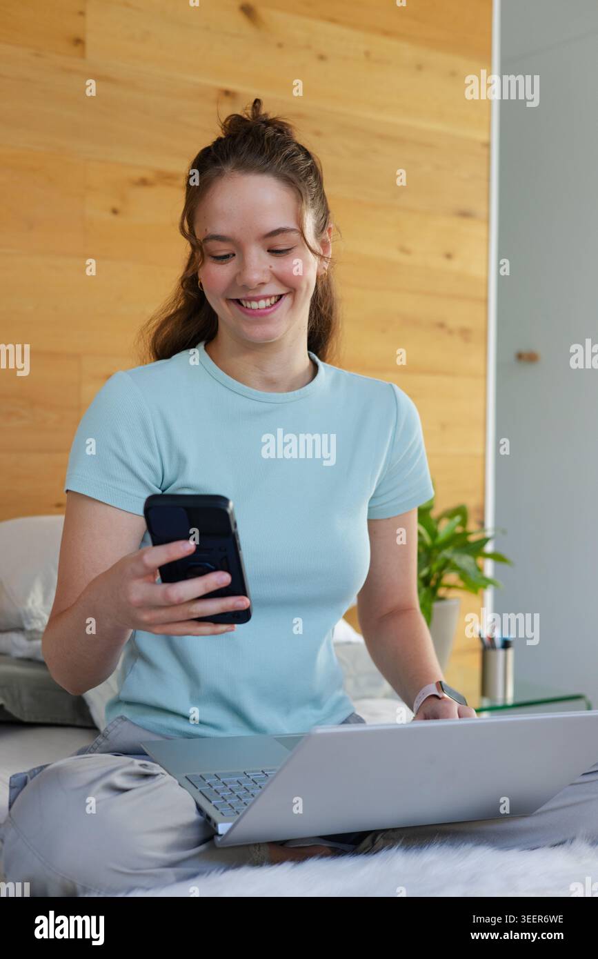 Woman sitting on bed, holding smartphone while using laptop, wearing light blue T shirt Stock Photo