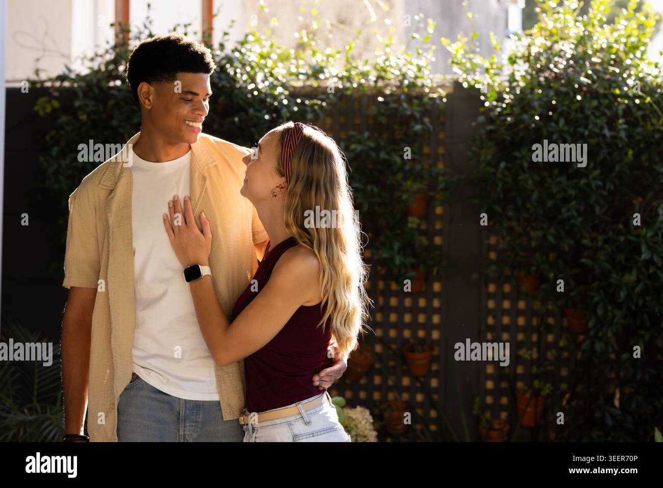 Diverse couple standing in sunlit backyard garden by wooden trellis, smiling, smartwatch visible Stock Photo