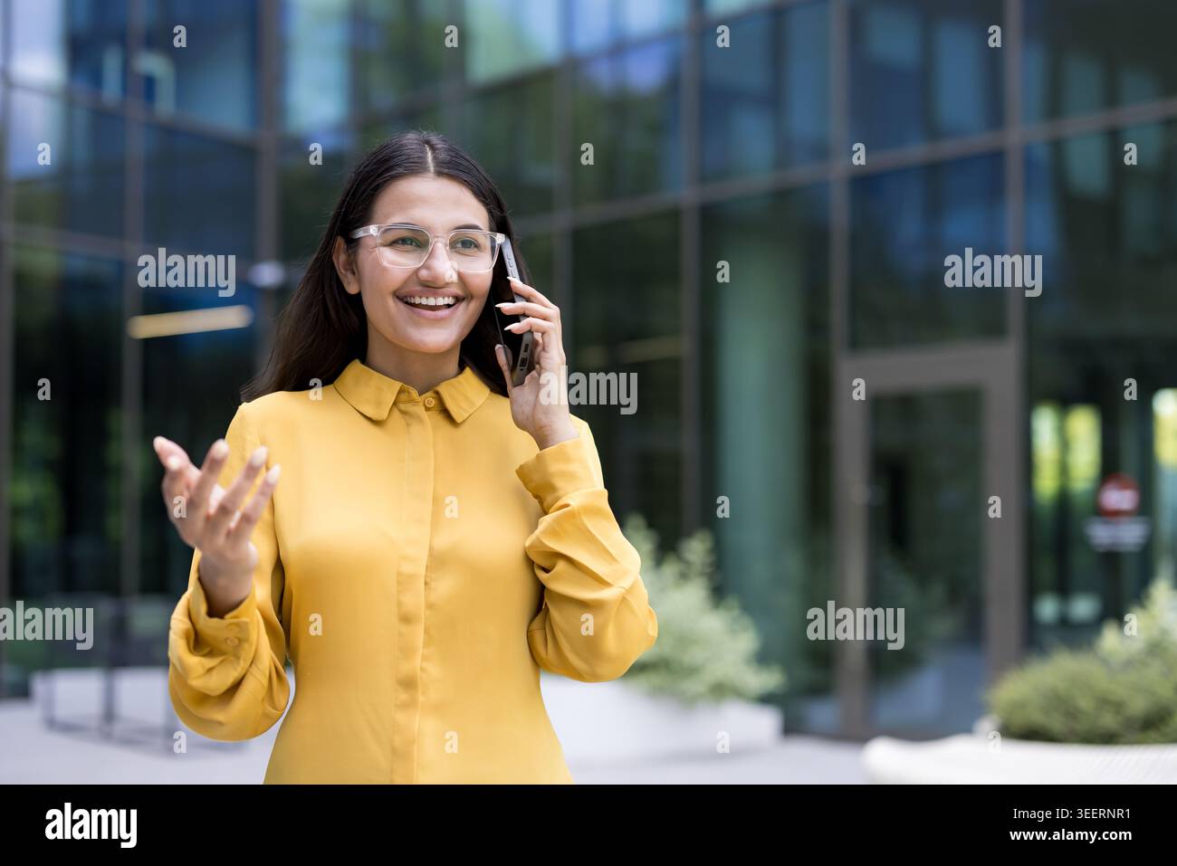 Indian businesswoman smiling confidently while having a professional conversation on her mobile phone outside a modern office building, representing corporate communication and success Stock Photo